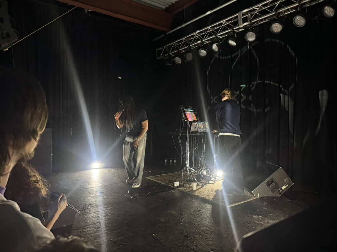 Rapper Nathaniel Ritchie and producer Parker Corey of the rap group By Storm performing at the Cat's Cradle Back Room. The photo is quite intimate, due to being taken from the front row of the venue. The room is quite dark, with only two flashing and strobing lights providing most of the illumination.