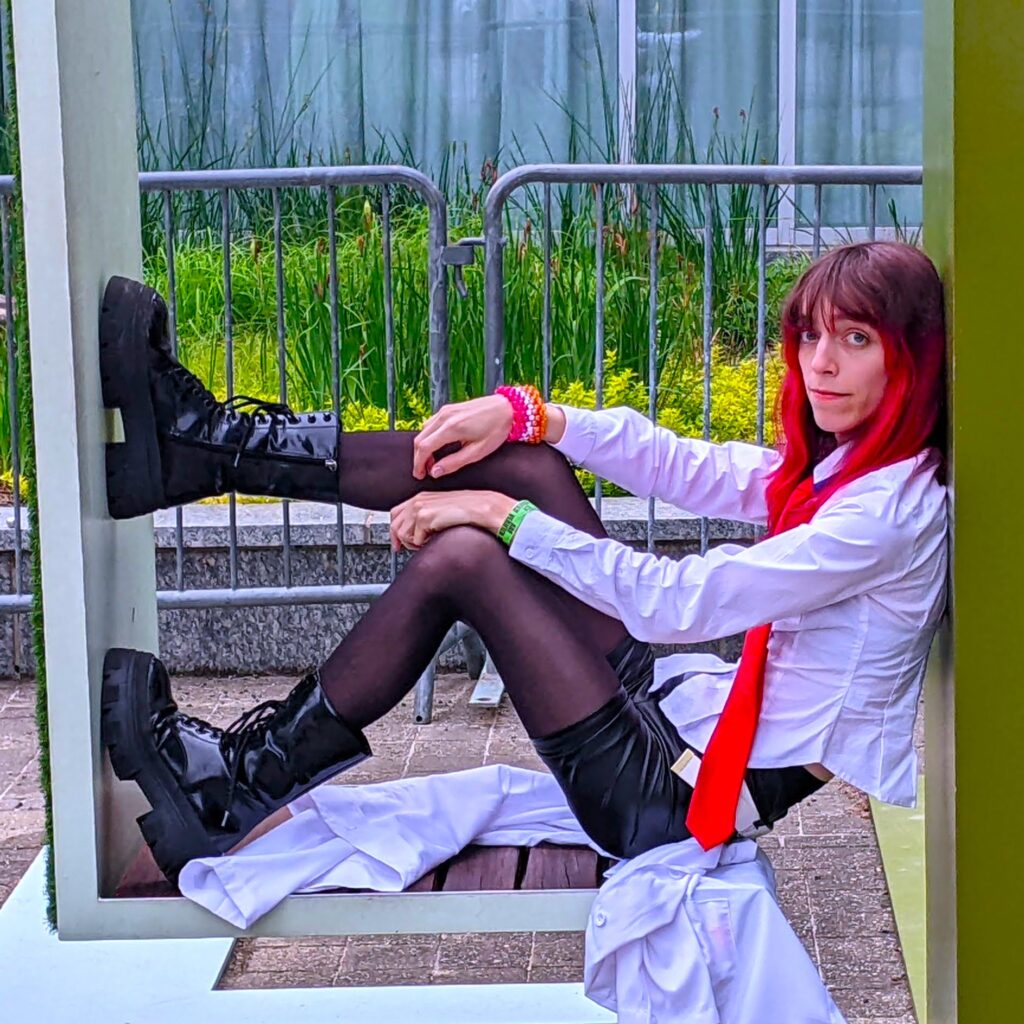A person with long, red-tipped hair wearing a white shirt, black leather skirt, and red tie, with a backdrop of green plants.