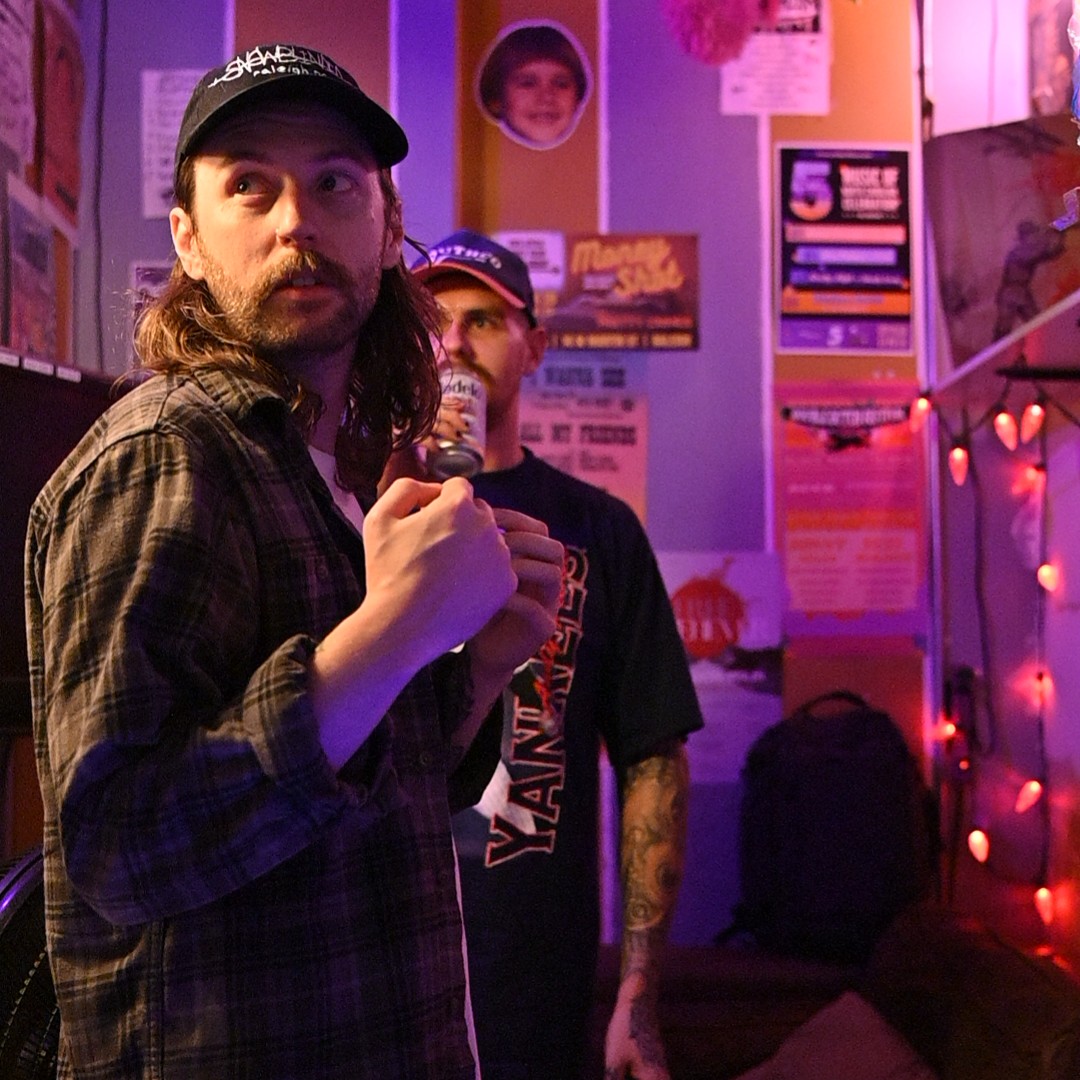 Cliff and Tom from the local NC band Wellspring taken in the Neptunes Parlor green room. They both look away from the camera due to it being more of a candid shot. Cliff, the vocalist, is in the foreground, while Tom, the guitarist, is in the background. Various alternative details adorn the walls, lending itself to a more D.I.Y. punk atmosphere