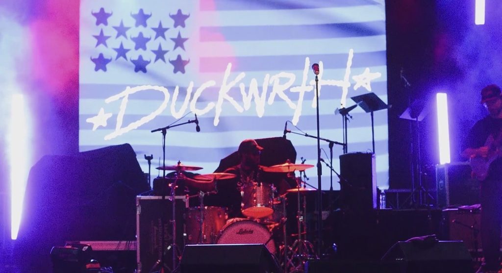 A man behind a set of drums, playing in front of a screen with the word "DUCKWRTH" shown.